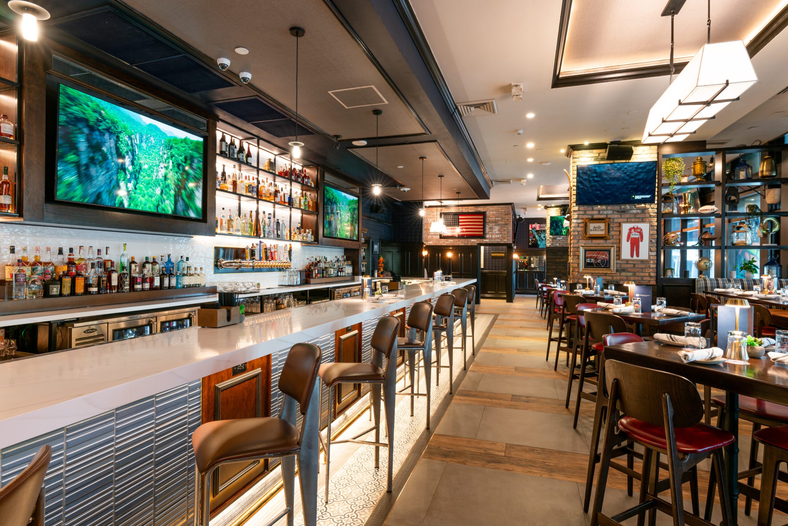 Modern bar interior with a long, sleek white countertop and stools. Shelves stocked with bottles and glasses line the wall behind the bar. Large TV screens display nature scenes. Tables are set with plates and glasses, and the lighting is warm and inviting.