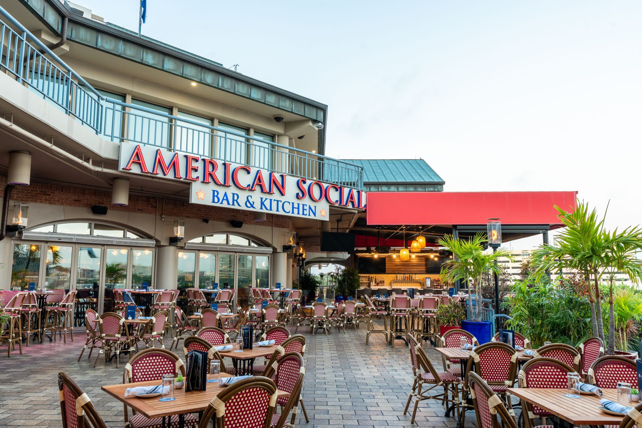The outdoor dining area of American Social Bar & Kitchen, known as Dining AmSo, boasts many empty tables on a brick patio. The restaurant features a partial roof, potted plants, and a large sign above the entrance.
