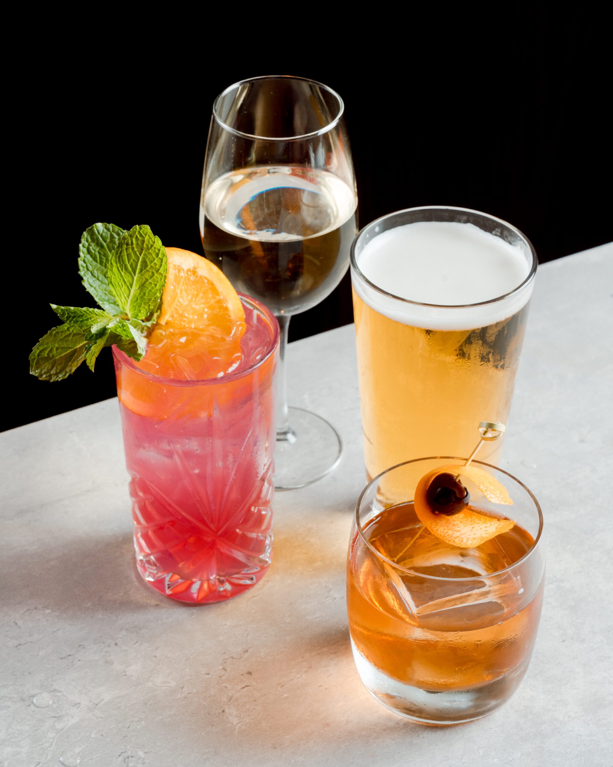 A variety of drinks on a light surface: a pink cocktail with mint and orange slice, a glass of white wine, a tall glass of beer, and a whiskey glass with an orange twist and cherry.