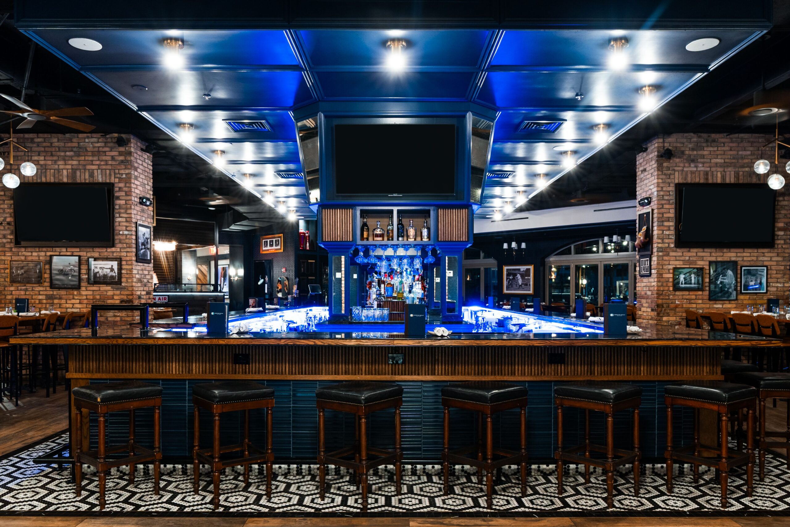 A modern Dining AmSo bar interior features a central bar counter surrounded by black barstools, illuminated shelves with bottles, overhead screens, brick walls, and patterned floor tiles. The space is empty and well-lit.