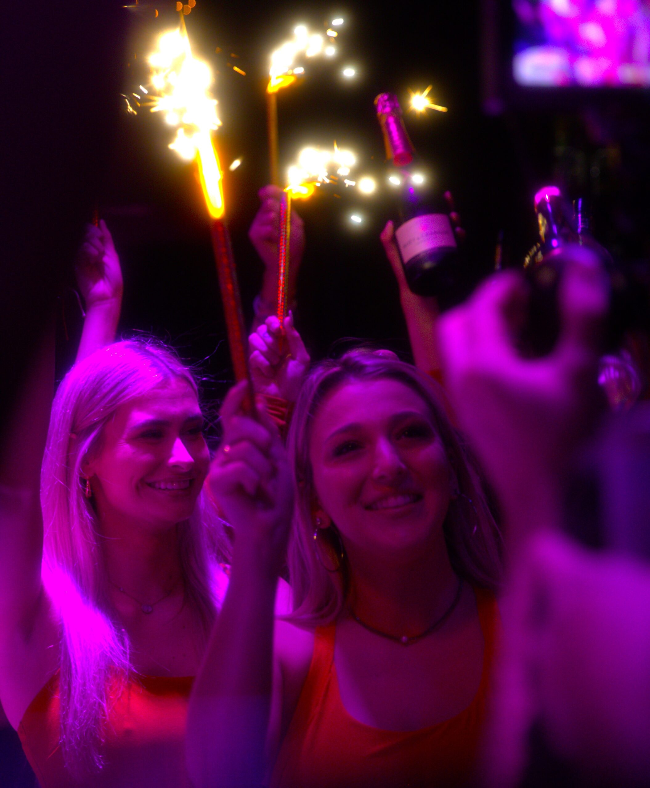 Two women smiling and holding sparklers in a crowd of people.