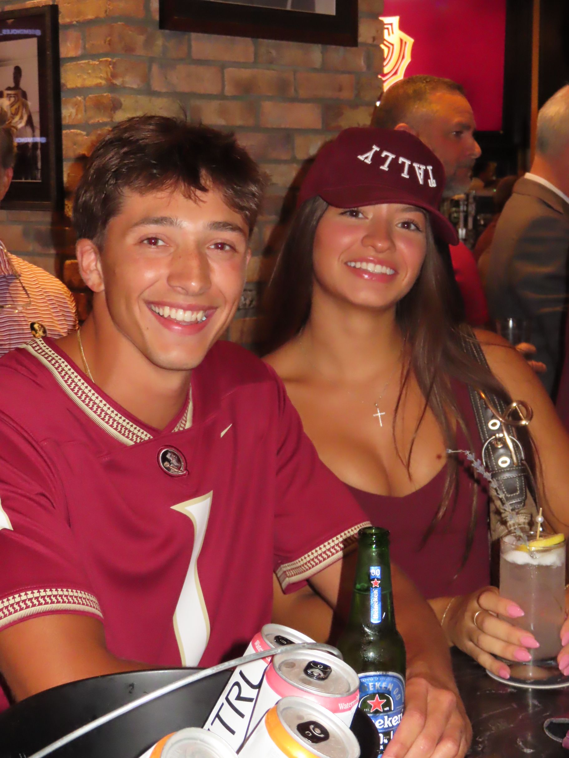 Two young adults in matching maroon sports jerseys smile at a bar. The woman wears a maroon hat, and both have drinks in front of them. A bucket of cans and bottles is on the table.