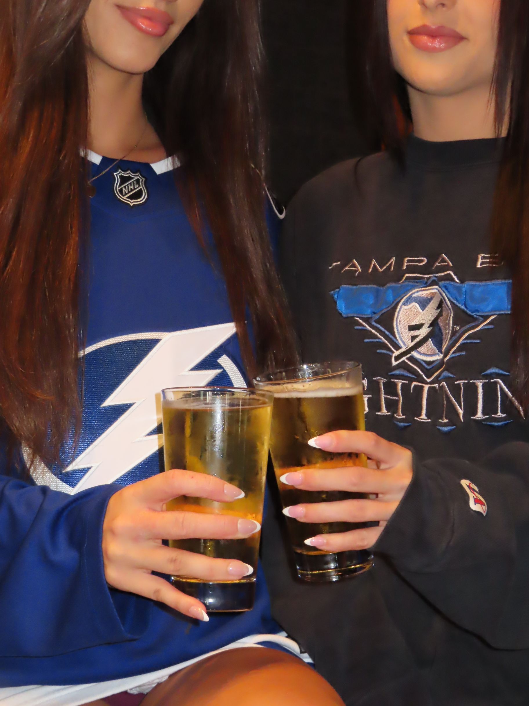 Two women wearing Tampa Bay Lightning shirts are holding and toasting with glasses of light-colored beer. Their faces are partially out of frame, and they have long brown hair.