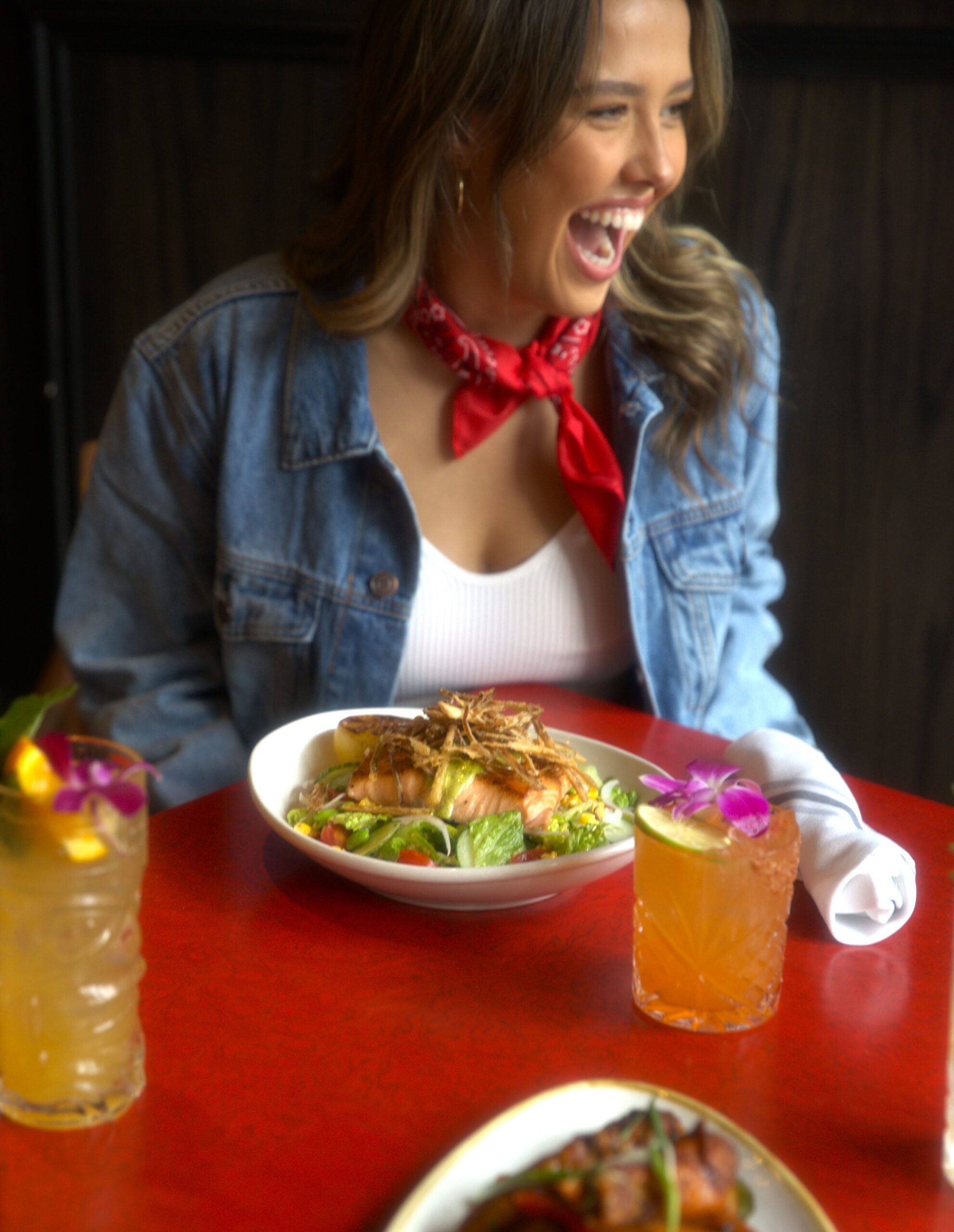 Laughing brown haired woman with food and drink in front of her.