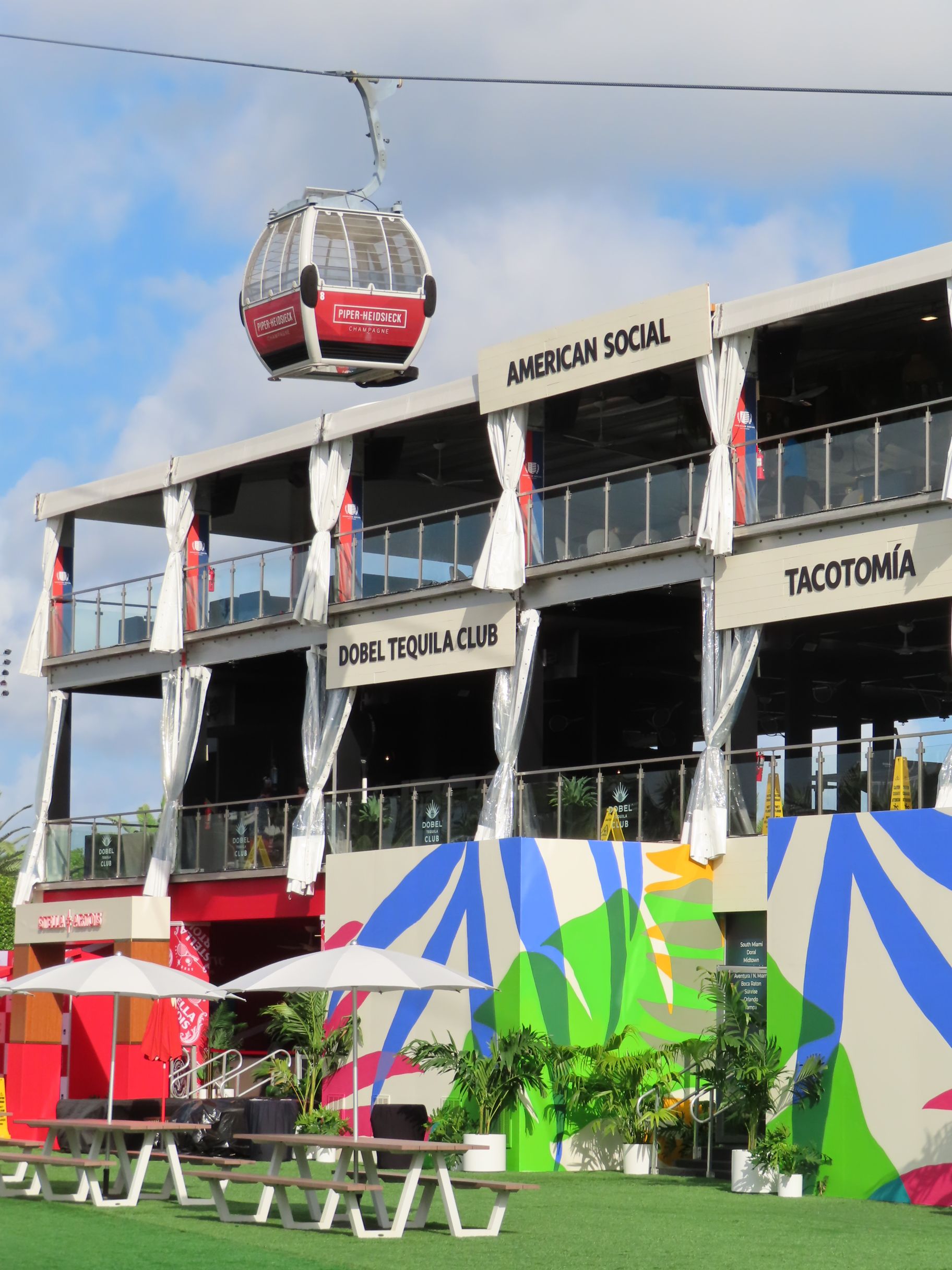 A gondola passes over a vibrant two-story building with signs for American Social, Dobel Tequila Club, and Tacotomía. Colorful murals, picnic tables, and umbrellas decorate the lawn below.