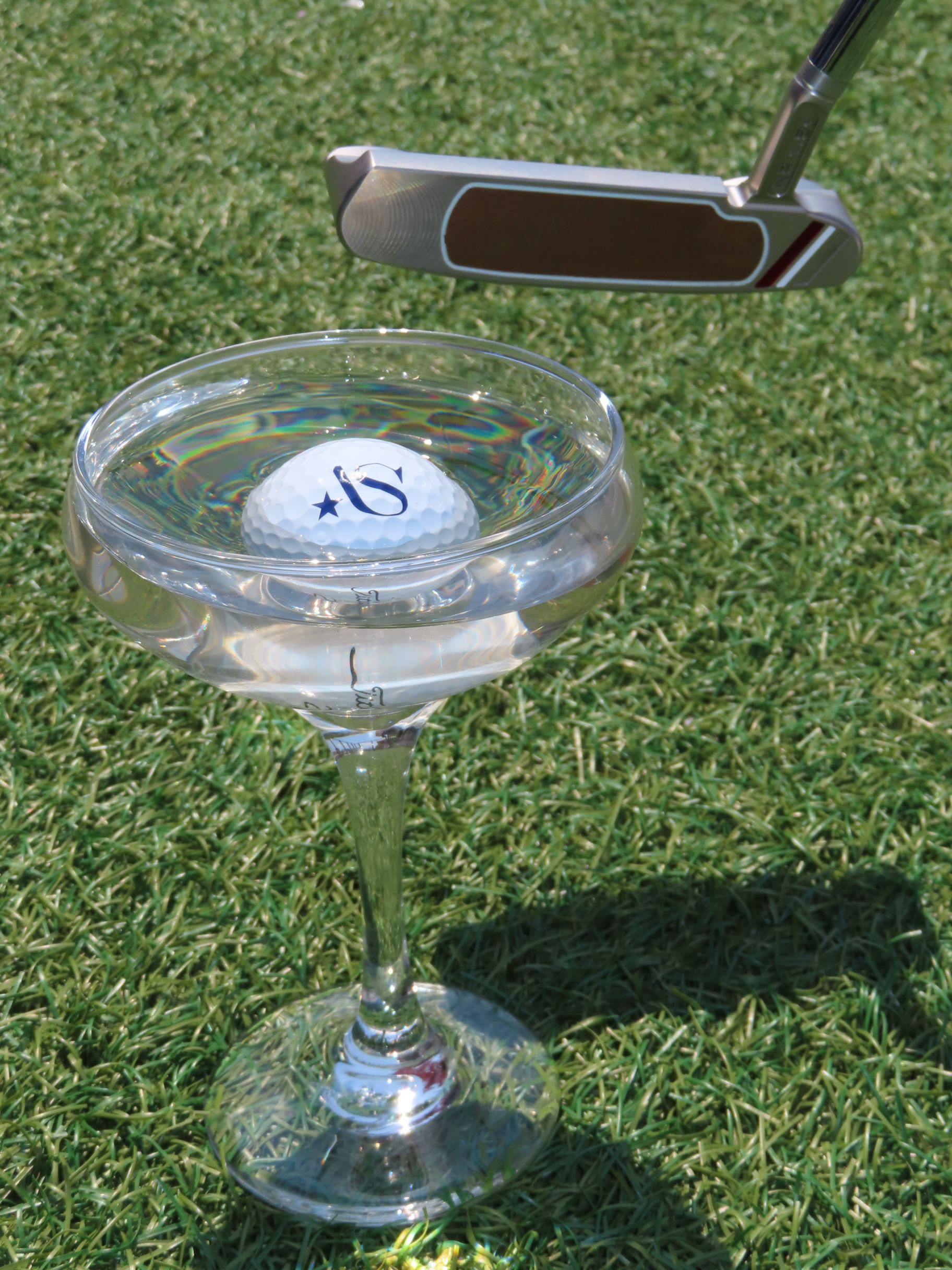 A golf ball floating in a cocktail glass filled with water sits on grass, with a golf putter poised above it as if to hit the ball.