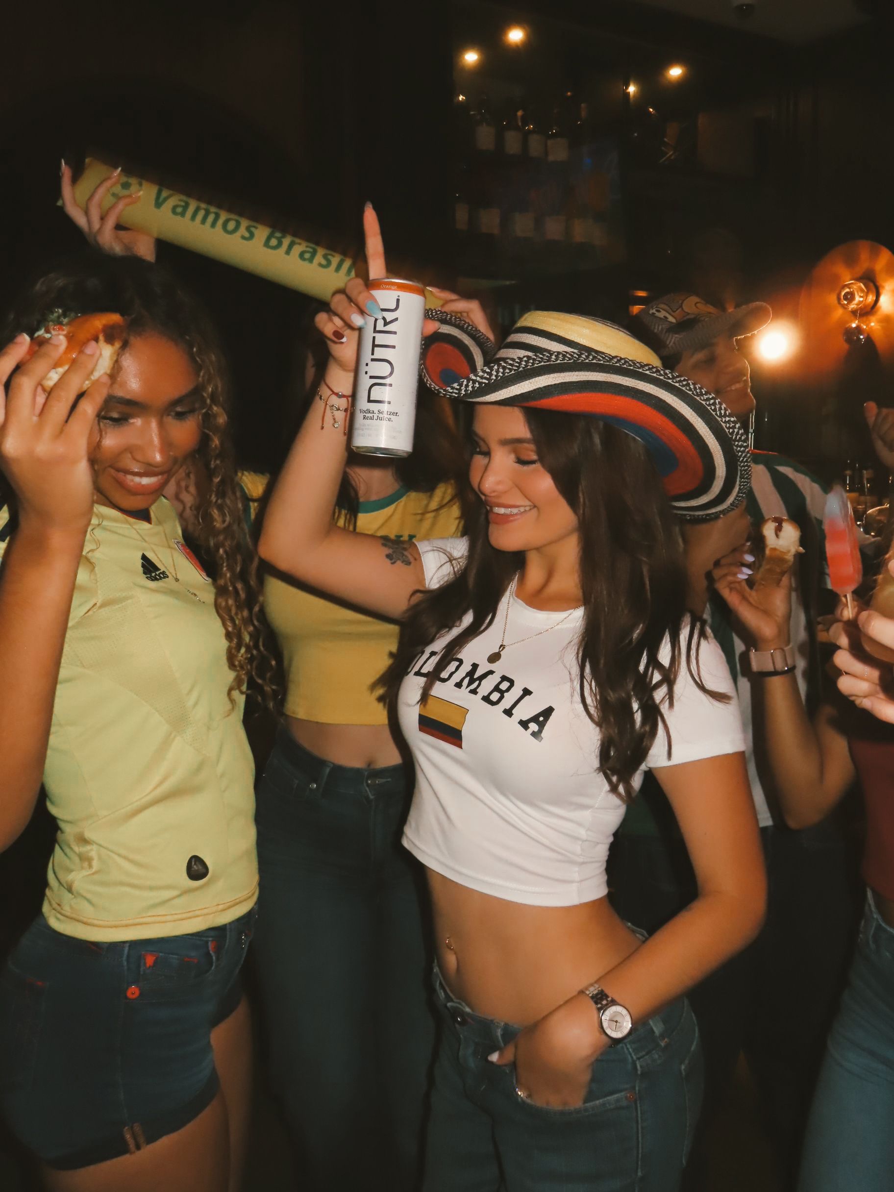 Two women dance at a lively party. One wears a yellow shirt and holds a snack, while the other, in a “Colombia” shirt and hat, smiles and holds a drink. Other people and festive decorations are visible in the background.