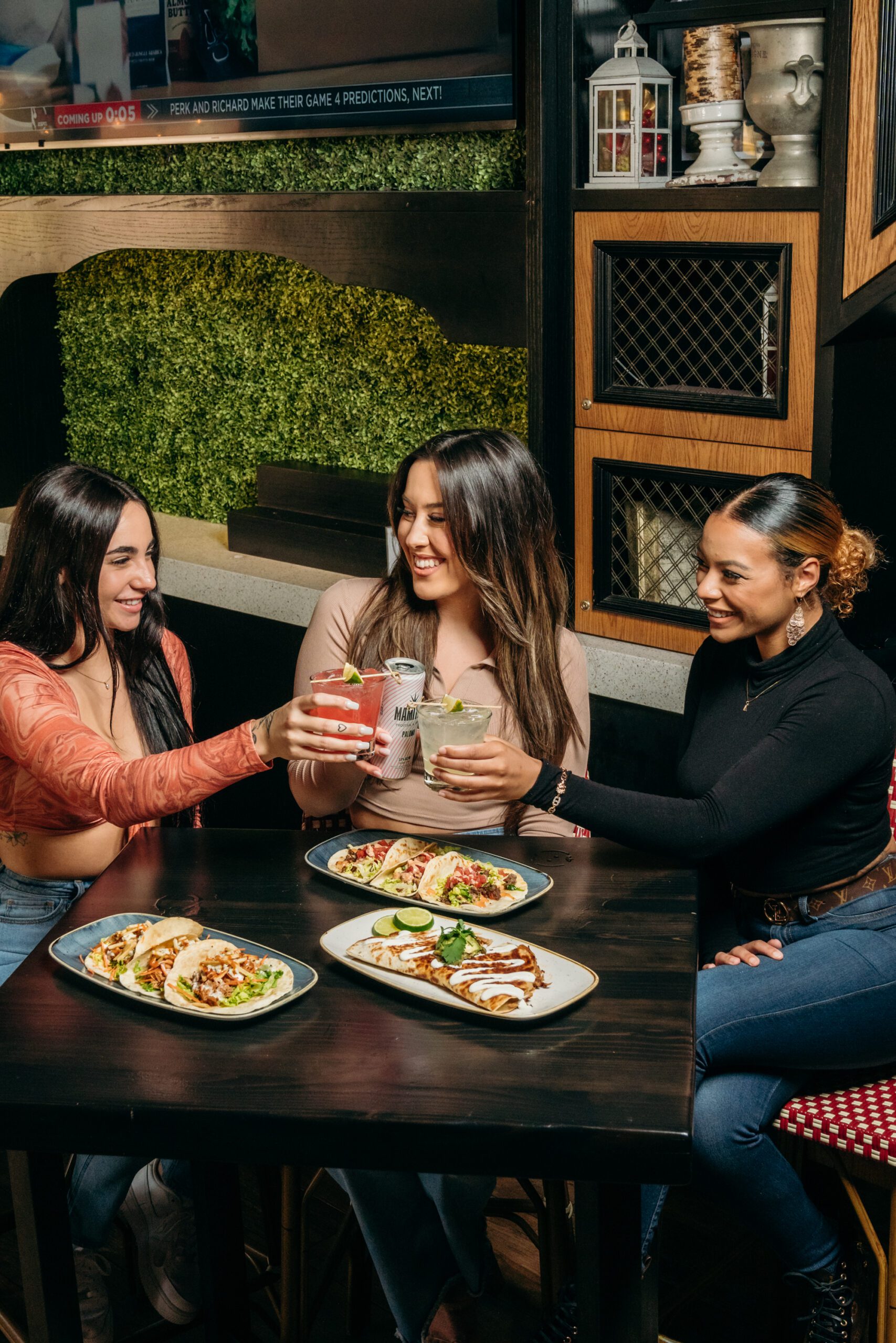 Three women are saying cheers with cocktails, sitting at a table with their dinner served.