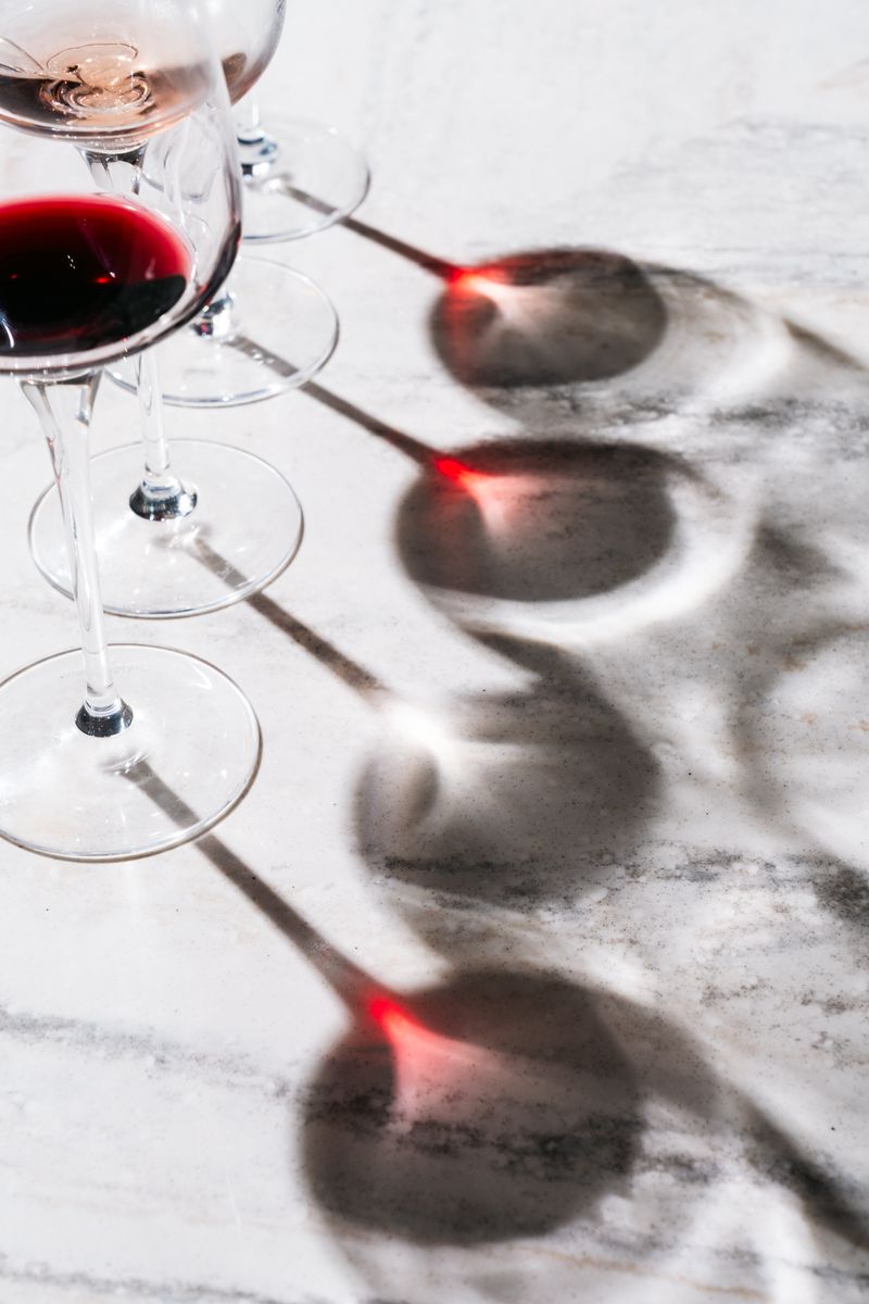 Four wine glasses, two with red wine and two empty, are arranged on a light marble surface. Sunlight casts dramatic shadows and red reflections—perfect for celebrating Wine Wednesdays in style.