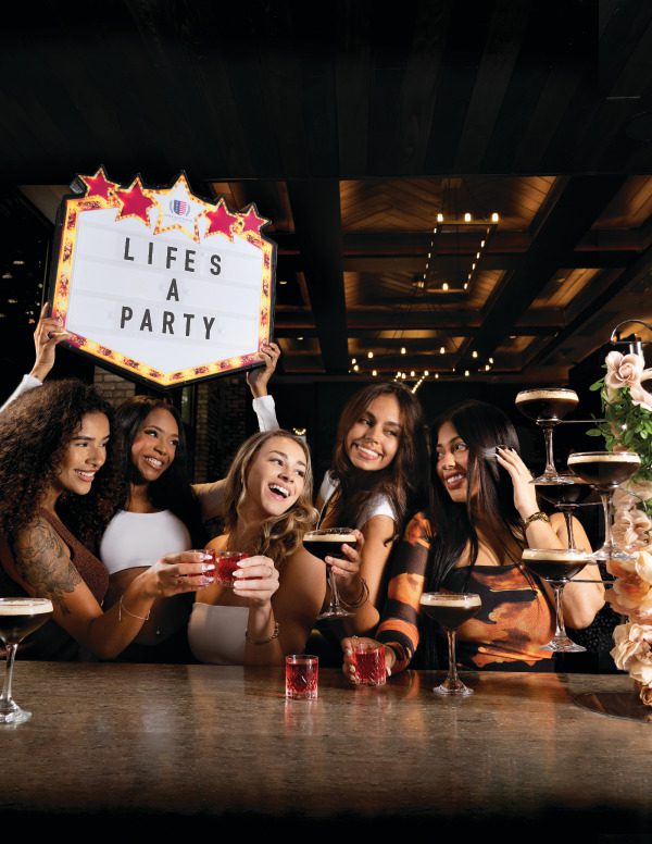 Five women at a bar holding drinks, one with a light-up 