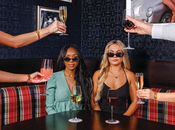 Two women in stylish outfits and sunglasses sit at a booth in a dark, trendy bar. Multiple hands holding various drinks extend toward them—it's the perfect scene for Wine Wednesdays. Framed photos decorate the wall behind them.