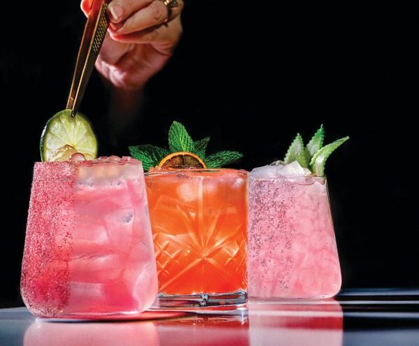 Three colorful cocktails with ice are shown; one is garnished with a lime slice by a hand holding tongs. Each drink is in a unique glass and decorated with fresh mint or citrus, set against a dark background.