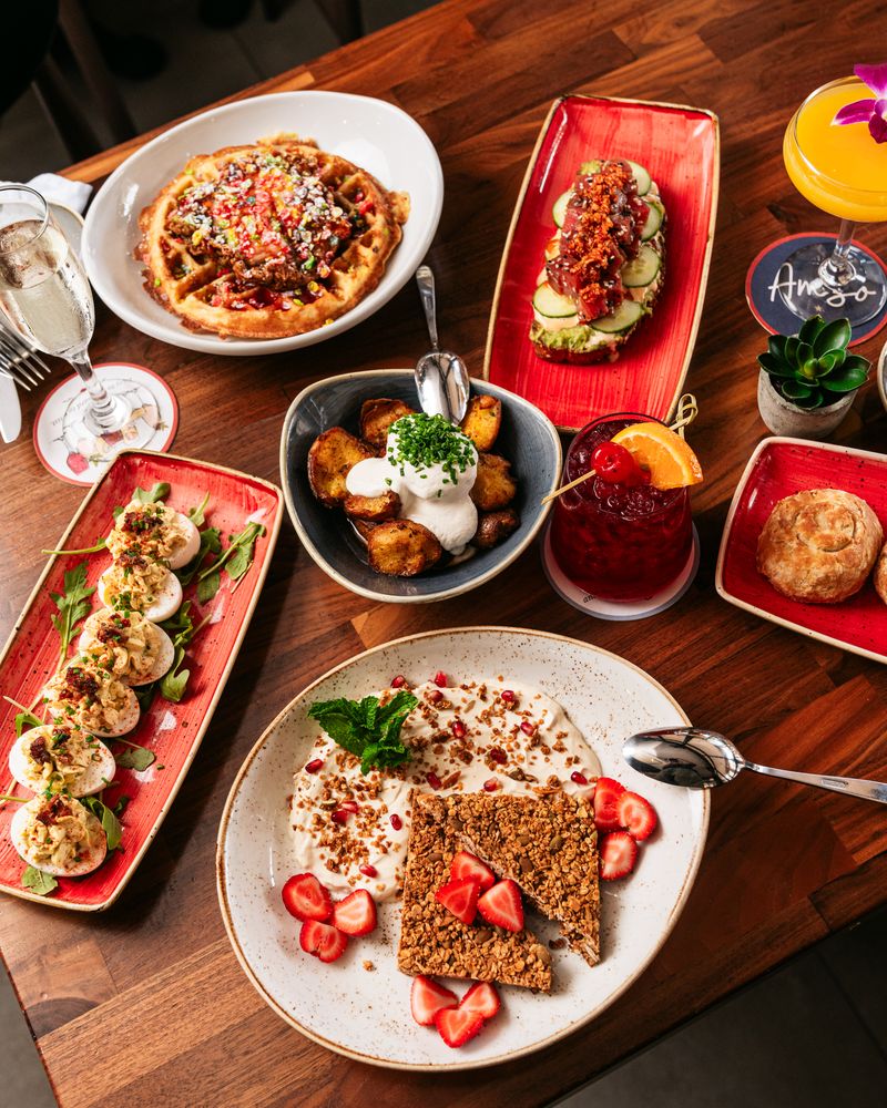 A wooden table set with assorted brunch dishes, including waffles, avocado toast, deviled eggs, crispy potatoes, yogurt with granola and strawberries, a biscuit, and colorful boozy drinks.