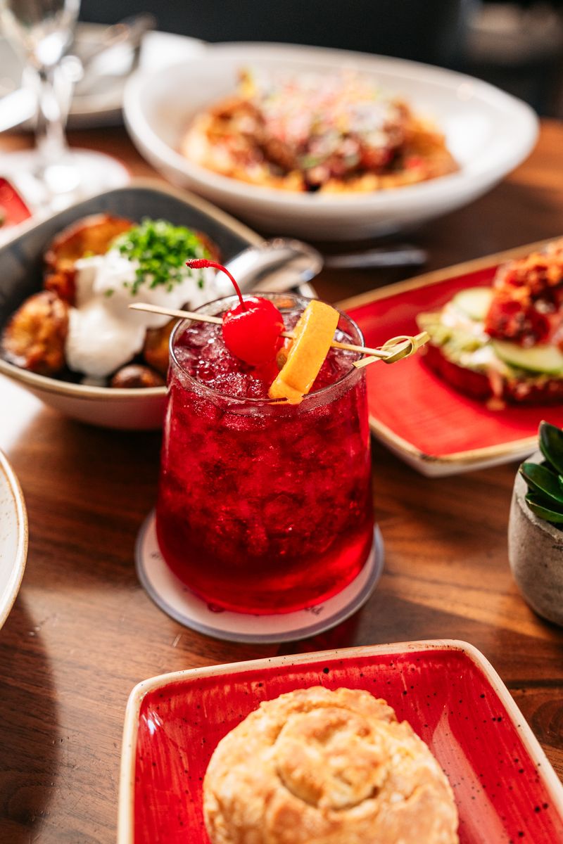 A vibrant, boozy red cocktail garnished with an orange slice and cherry sits on a wooden table, surrounded by plates of food including biscuits, roasted potatoes with sour cream, and a pasta dish in the background.
