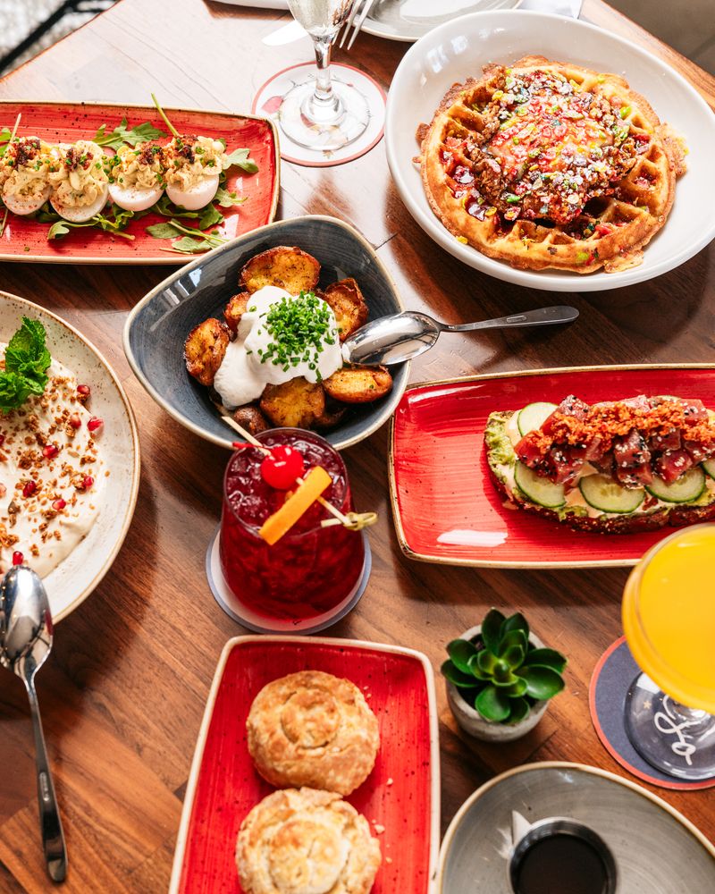 A wooden table set with deviled eggs, waffles with syrup and nuts, crispy potatoes with sour cream, avocado toast with bacon, a boozy red cocktail, an orange drink, biscuits, and a small potted plant.