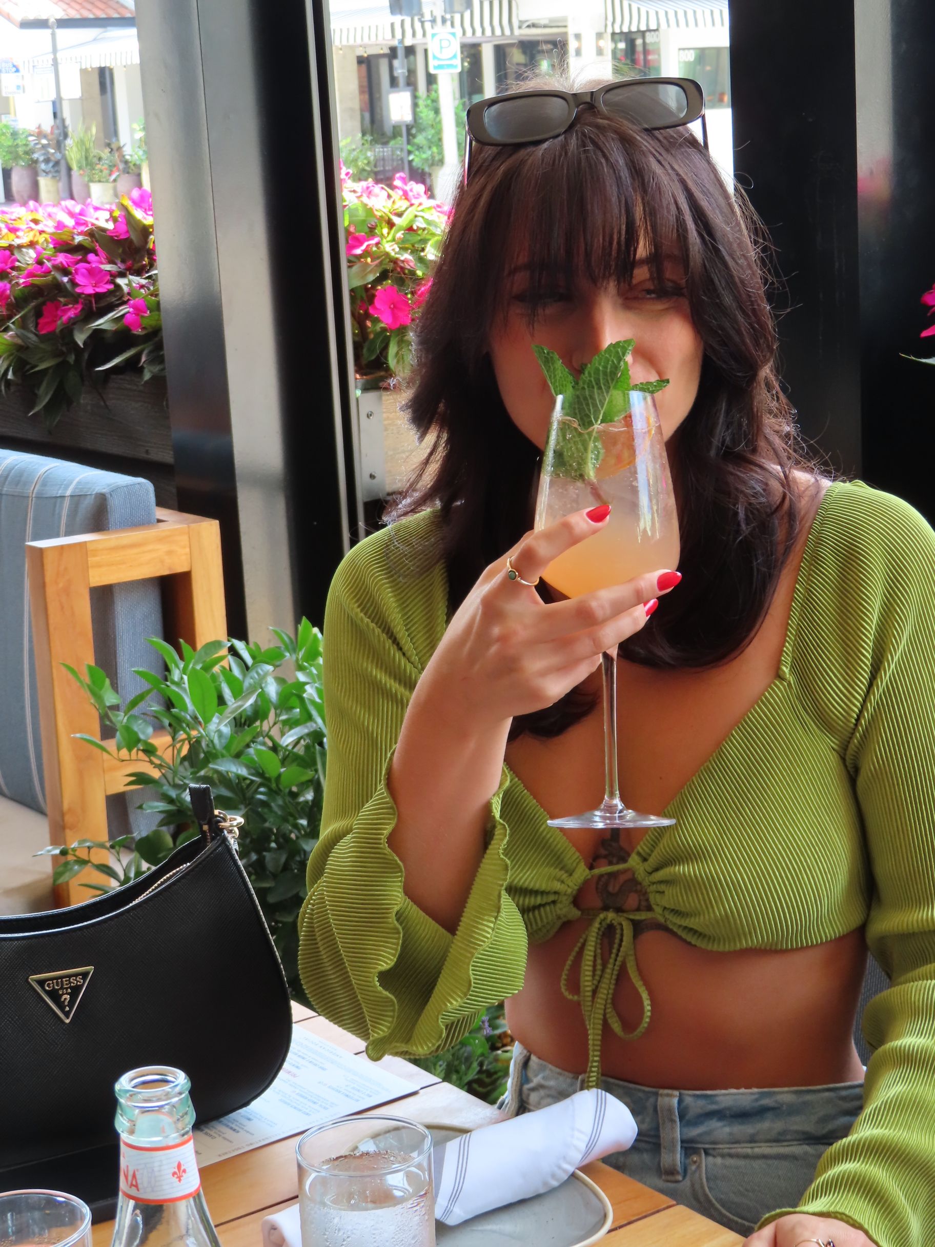 A woman with dark hair and sunglasses on her head sits at an outdoor restaurant, smiling and holding a boozy cocktail garnished with mint. She wears a green cropped top, red nail polish, and has a black Guess purse beside her.