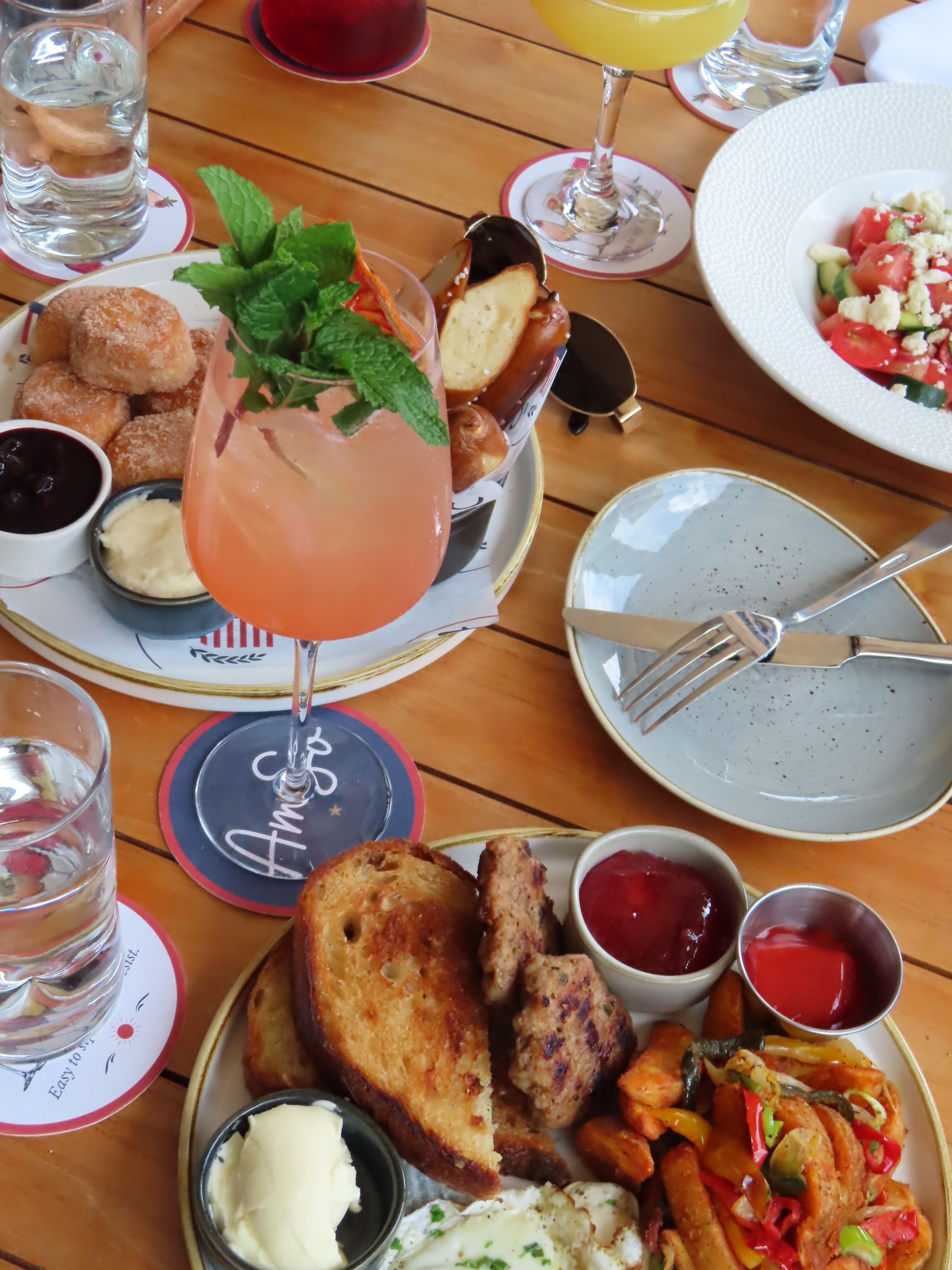 A table set for brunch with assorted dishes, including toast, grilled vegetables, dips, potato bites, and a salad. Boozy, colorful cocktails garnished with mint sit alongside glasses of water on the wooden table.
