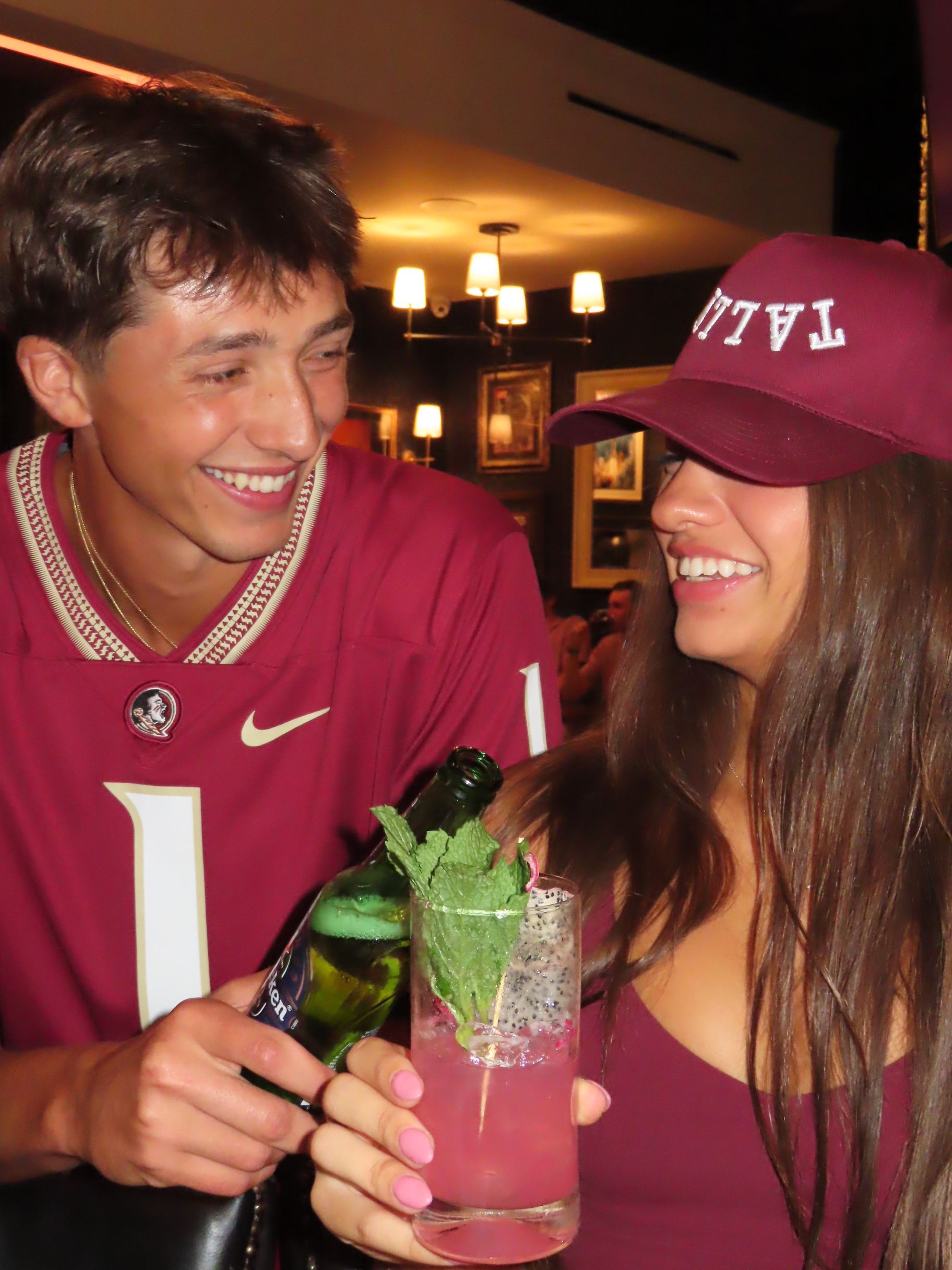 A young man and woman in matching maroon outfits smile at each other in a bar, their style subtly nodding to FSU football spirit. The woman holds a pink cocktail with mint, while the man pours a drink, both appearing cheerful and relaxed.