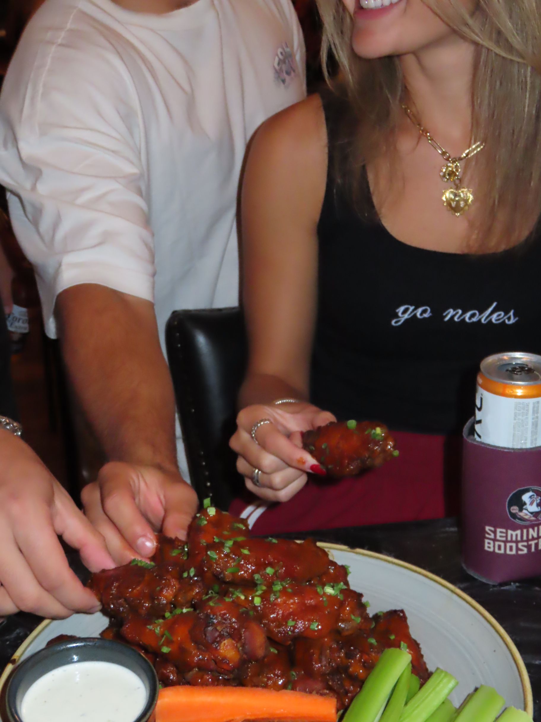 Two people sit together, smiling and sharing saucy chicken wings with carrot and celery sticks as they celebrate an exciting FSU football game day. A drink in a can and a cup labeled “Seminole Booster” are on the table beside a small bowl of ranch dressing.