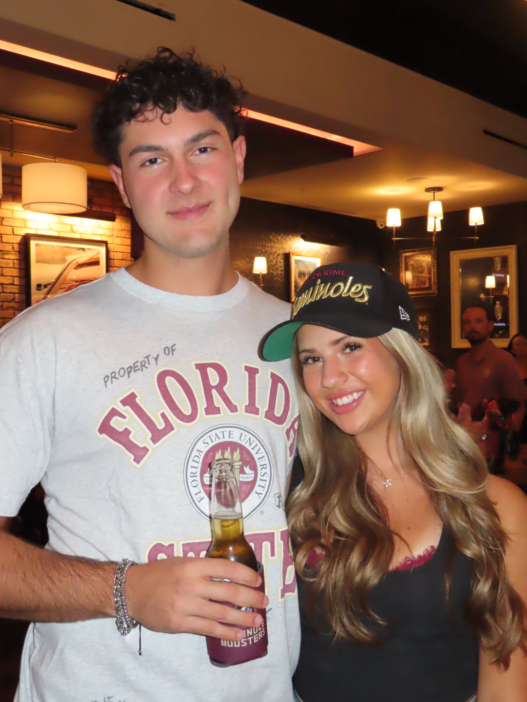 A smiling young man in a Florida State shirt—clearly an FSU football fan—holds a beer, standing next to a smiling young woman with long hair wearing a black Orioles cap. They are inside a warmly lit restaurant or bar.