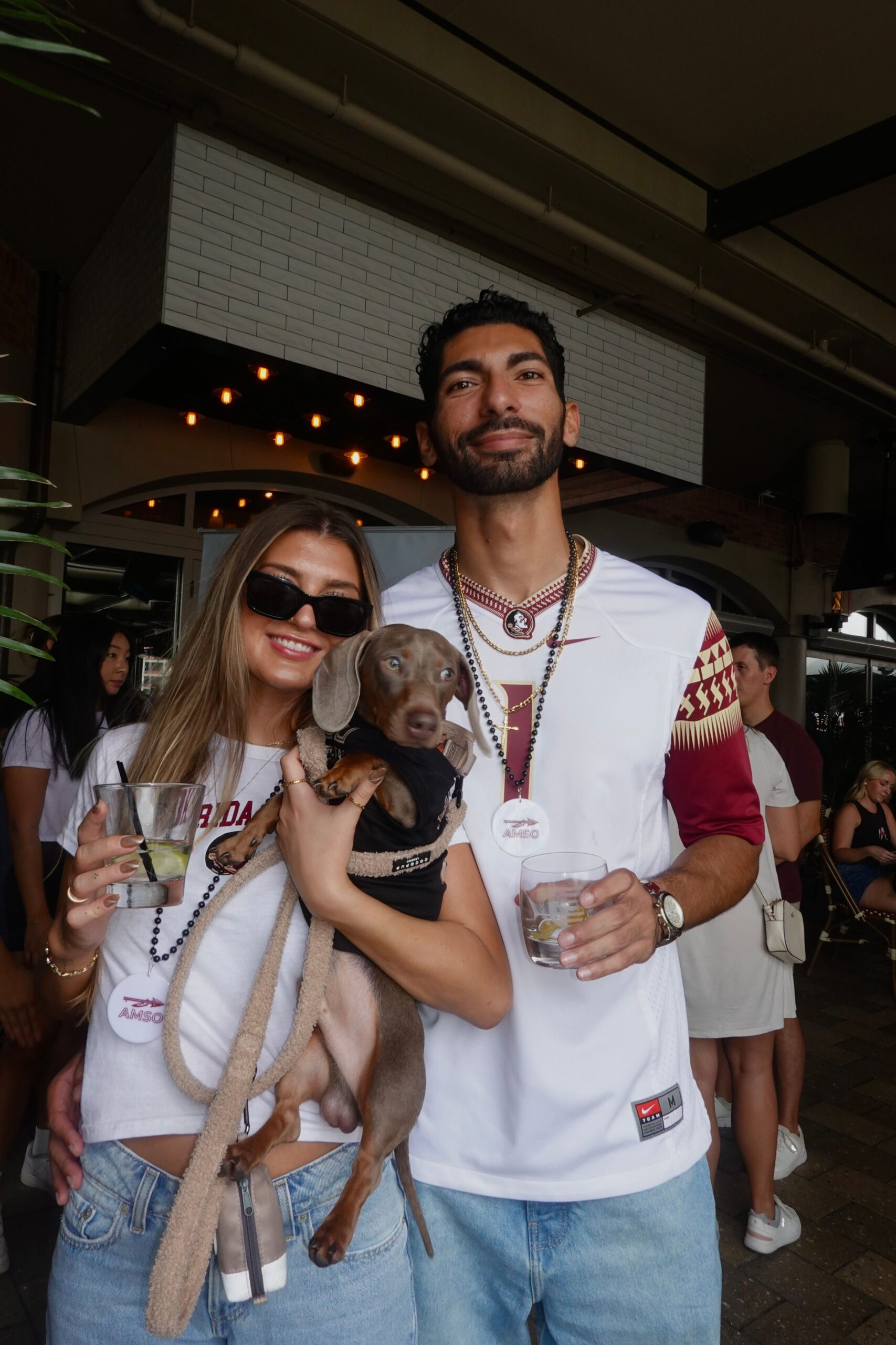 A smiling woman and man stand together at an outdoor event, each holding a drink. The woman is holding a small dog. Both wear casual clothes, beads, and sunglasses—showing off their fun spirit at what appears to be an FSU football gathering. Other people are visible in the background.