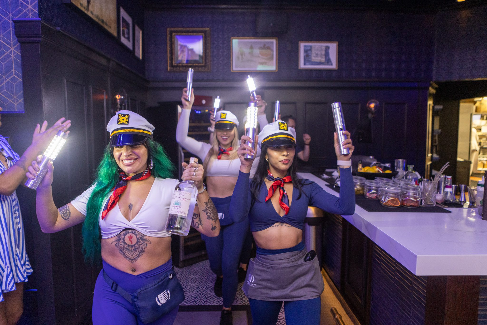 Three people dressed in themed sailor outfits hold glowing bottles and walk joyfully through a dimly lit room, reveling in the spirit of Halloween events. They wear hats and red neckerchiefs, amidst dark walls, framed pictures, and a bar counter with various items.