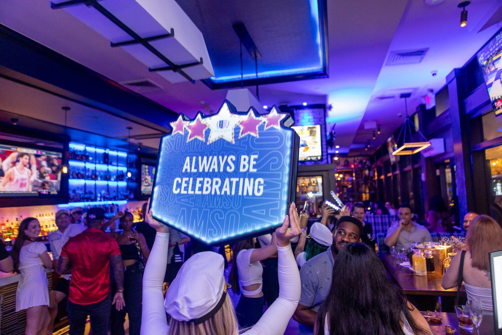 People enjoy a lively night at a bar with neon blue lighting, reminiscent of vibrant nautical nights. A person in the foreground holds a sign reading 