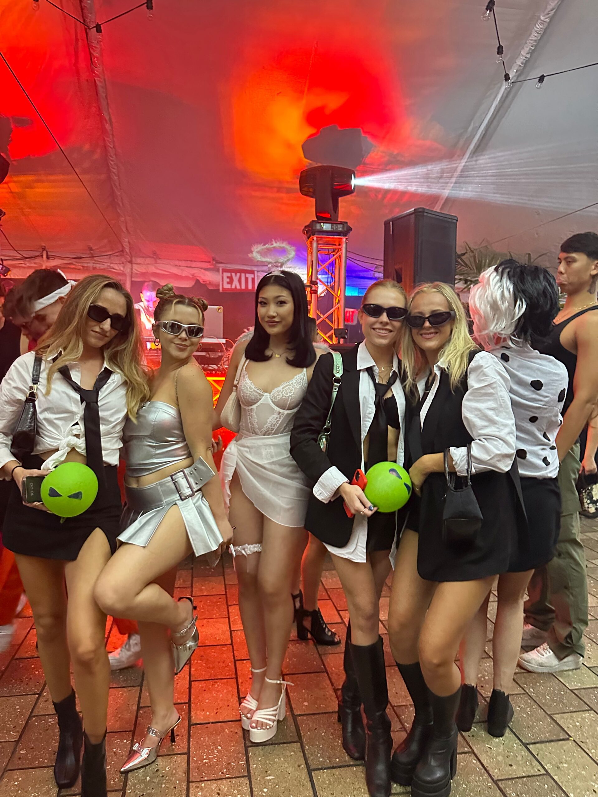 A group of six people stand under a tent with colorful lights, perfectly embodying the spirit of Halloween events. They are dressed in costumes, including a futuristic silver outfit, a white lace dress, and black-and-white spy attire with sunglasses. Some hold round green bags.
