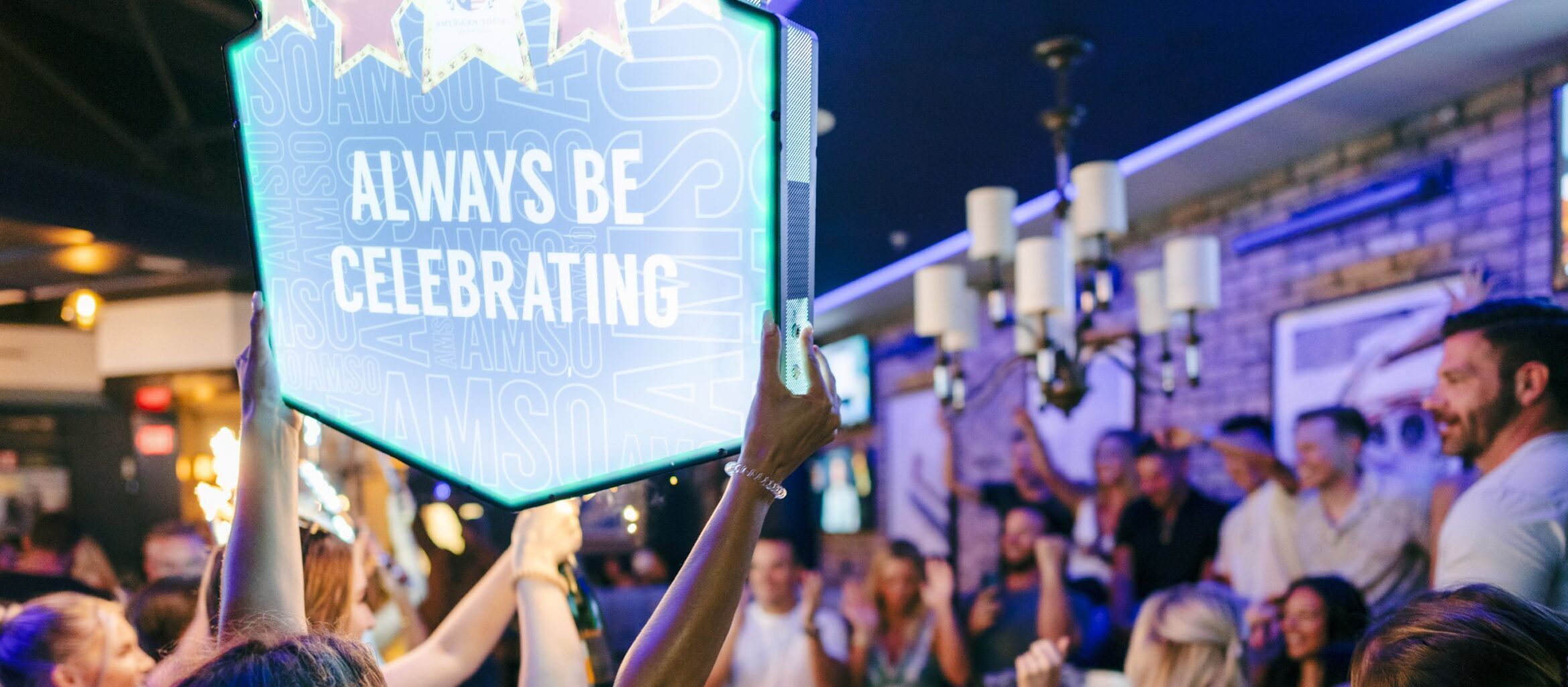 A lively group of people in a bar are energetically celebrating. One person in the foreground holds up a neon sign saying