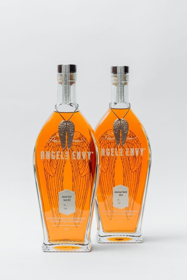 Two bottles of Angel's Envy bourbon with orange-brown liquid inside, perfect for Valentine's Day. The clear bottles feature wing illustrations and silver caps, standing on a white background.