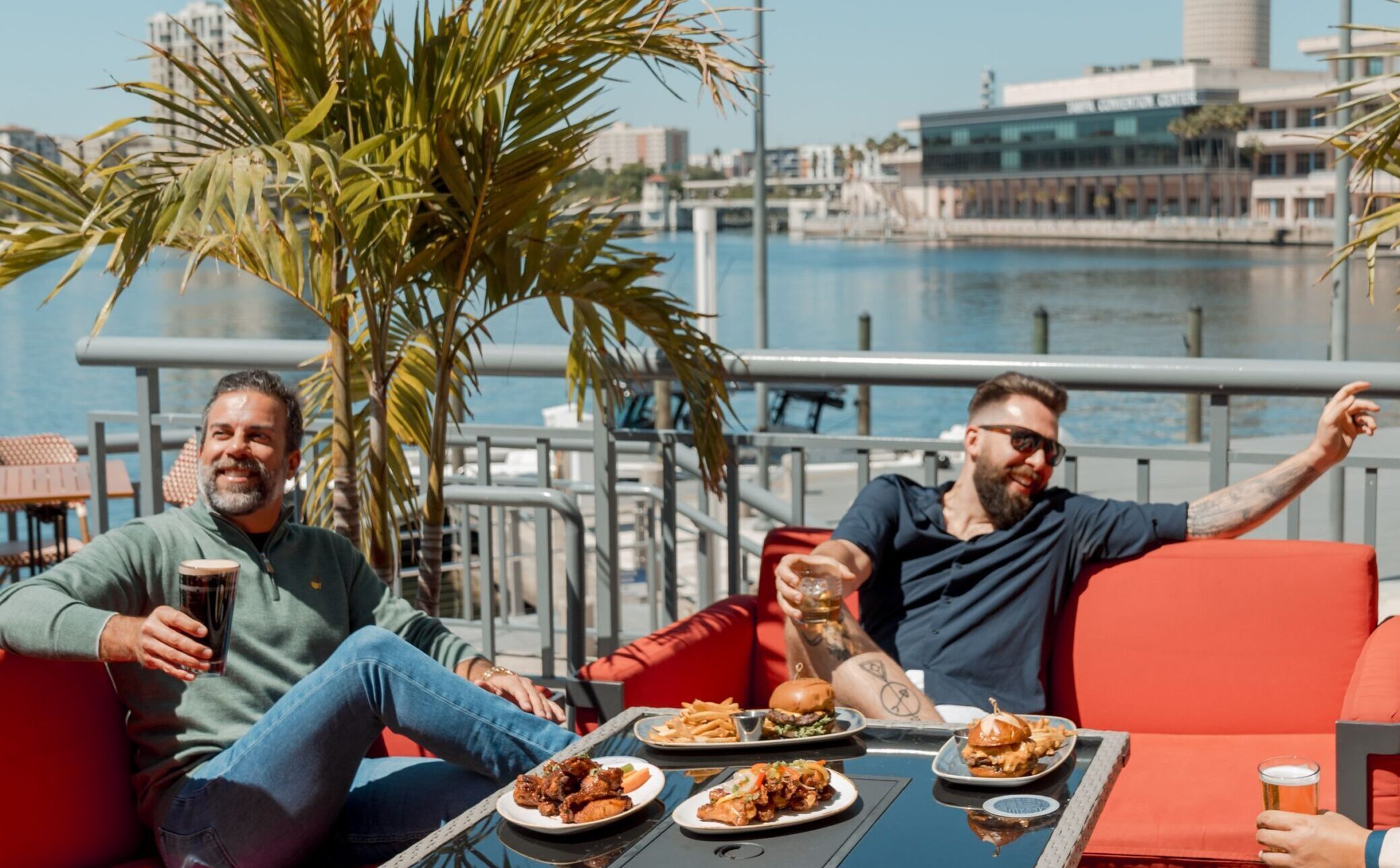 Three men sit on a bright red outdoor sofa by the water, laughing and enjoying drinks and plates of food on a sunny Valentine’s Day, surrounded by palm trees and waterfront buildings.