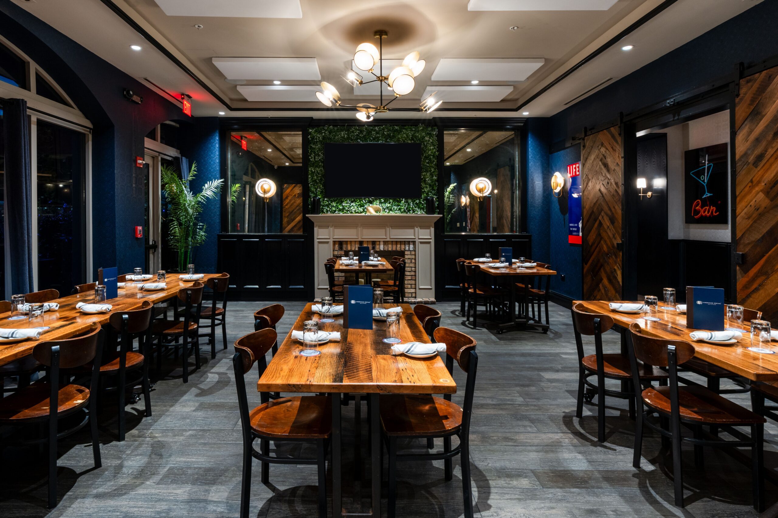 A stylish restaurant dining room with wooden tables and chairs, blue menus, place settings, modern lighting, and dark blue walls. Large windows and decorative greenery enhance the cozy atmosphere—perfect for private events in Tampa.