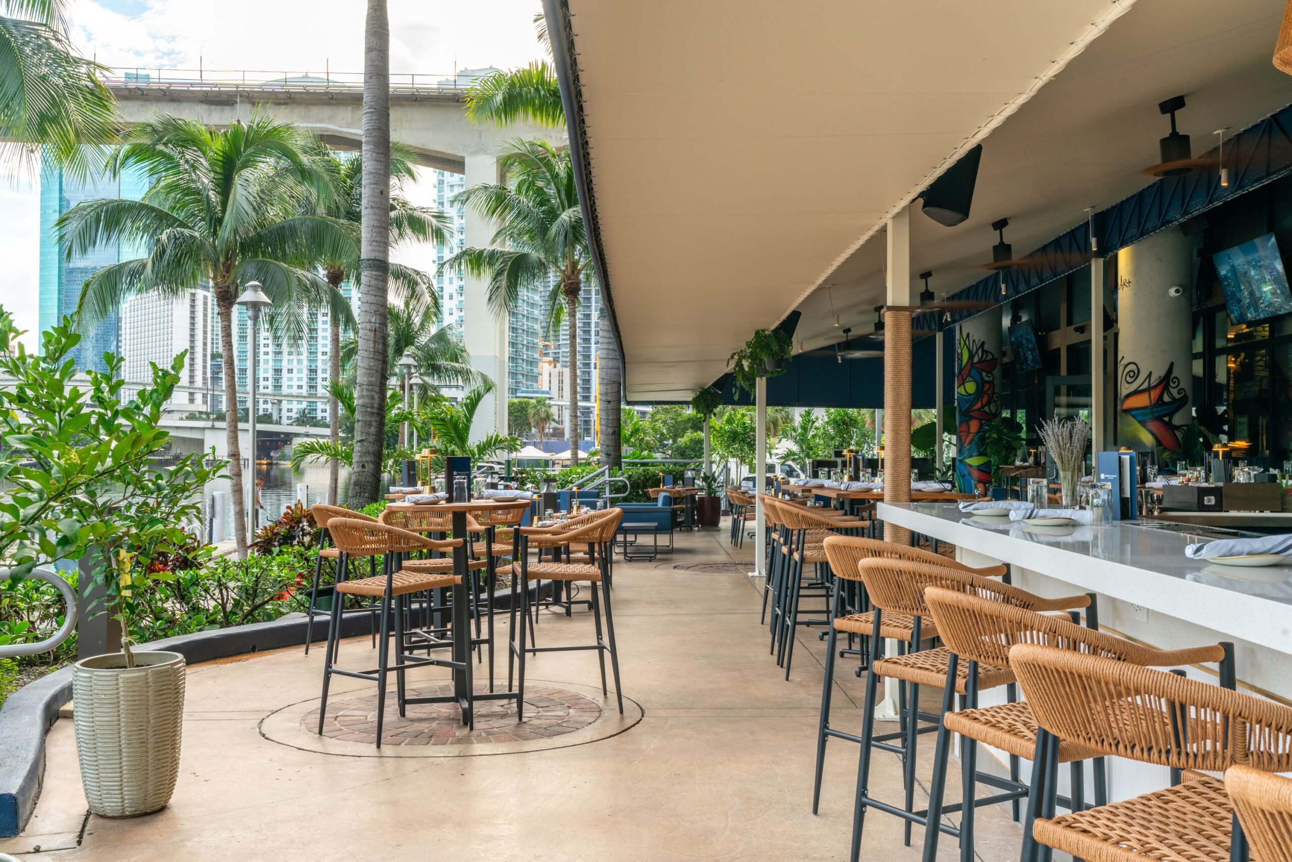 An outdoor restaurant patio with wicker chairs and high tables, surrounded by tropical plants and palm trees, overlooks a cityscape—an ideal setting for Private Events against the backdrop of tall buildings and an elevated roadway.