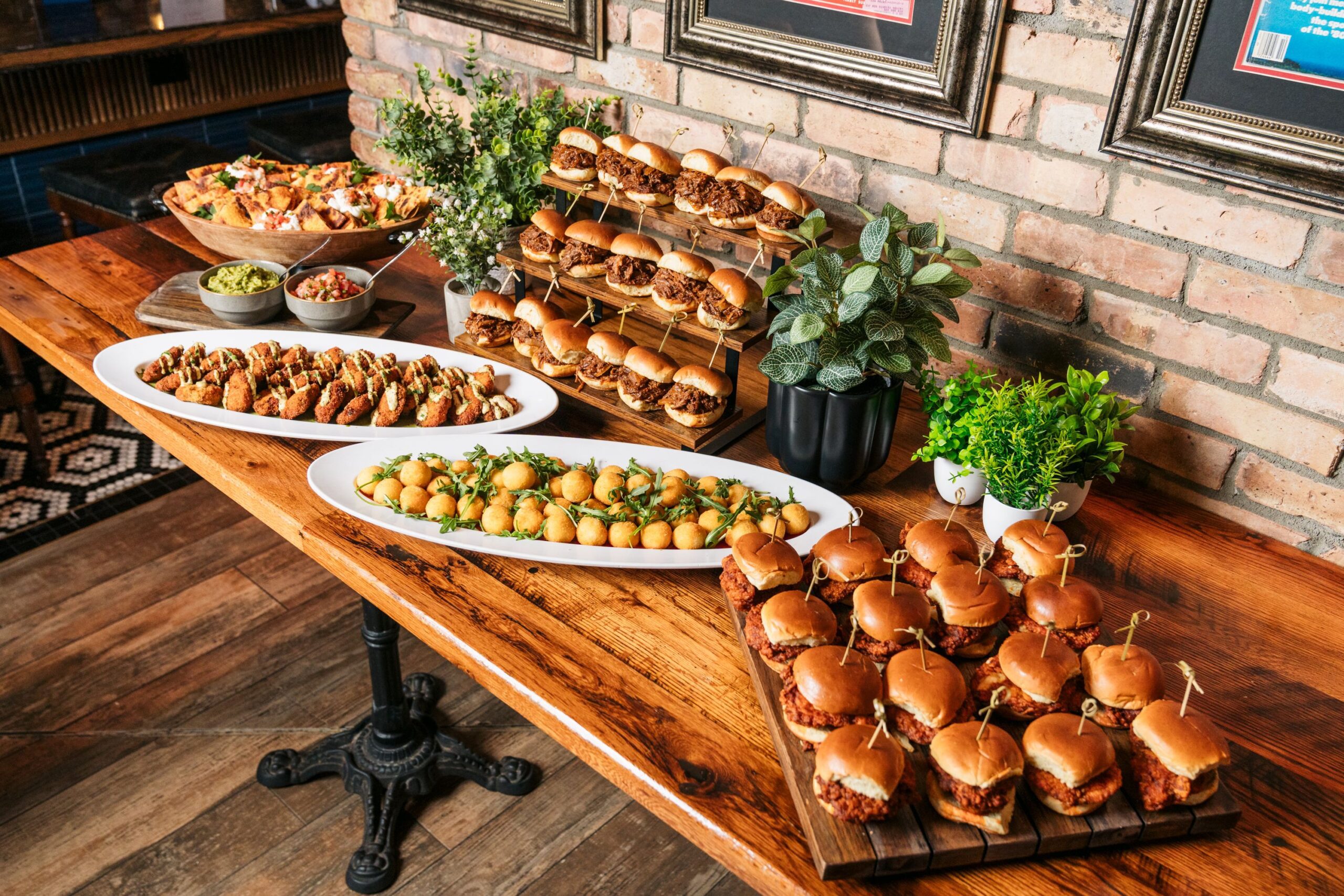 A wooden table set with platters of mini burgers, fried appetizers, and dipping sauces, surrounded by green plants against a brick wall—perfect for hosting private events in Tampa.