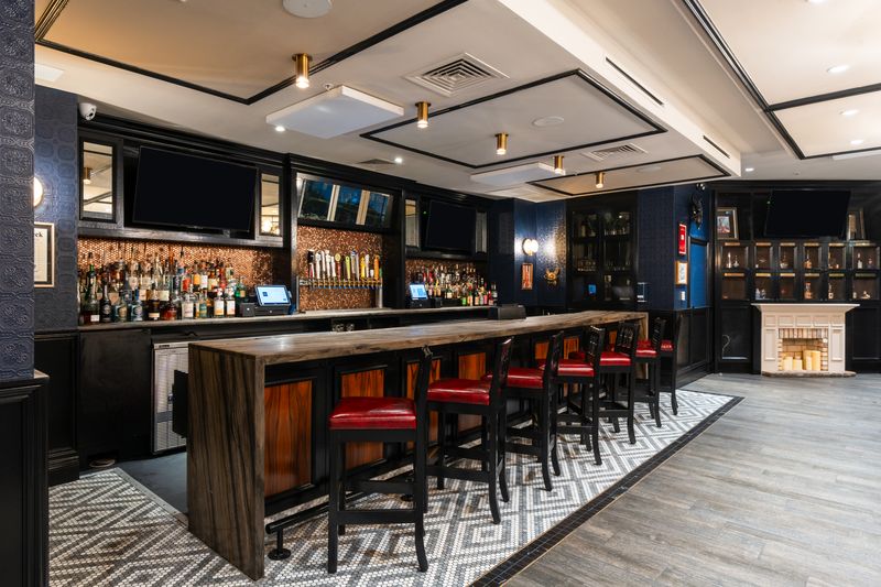 Modern bar with a long wooden counter, red cushioned stools, and shelves stocked with bottles behind the bar. Patterned tile flooring, dark accents, and warm lighting make it an ideal spot for Private Events Tampa. No people are present.