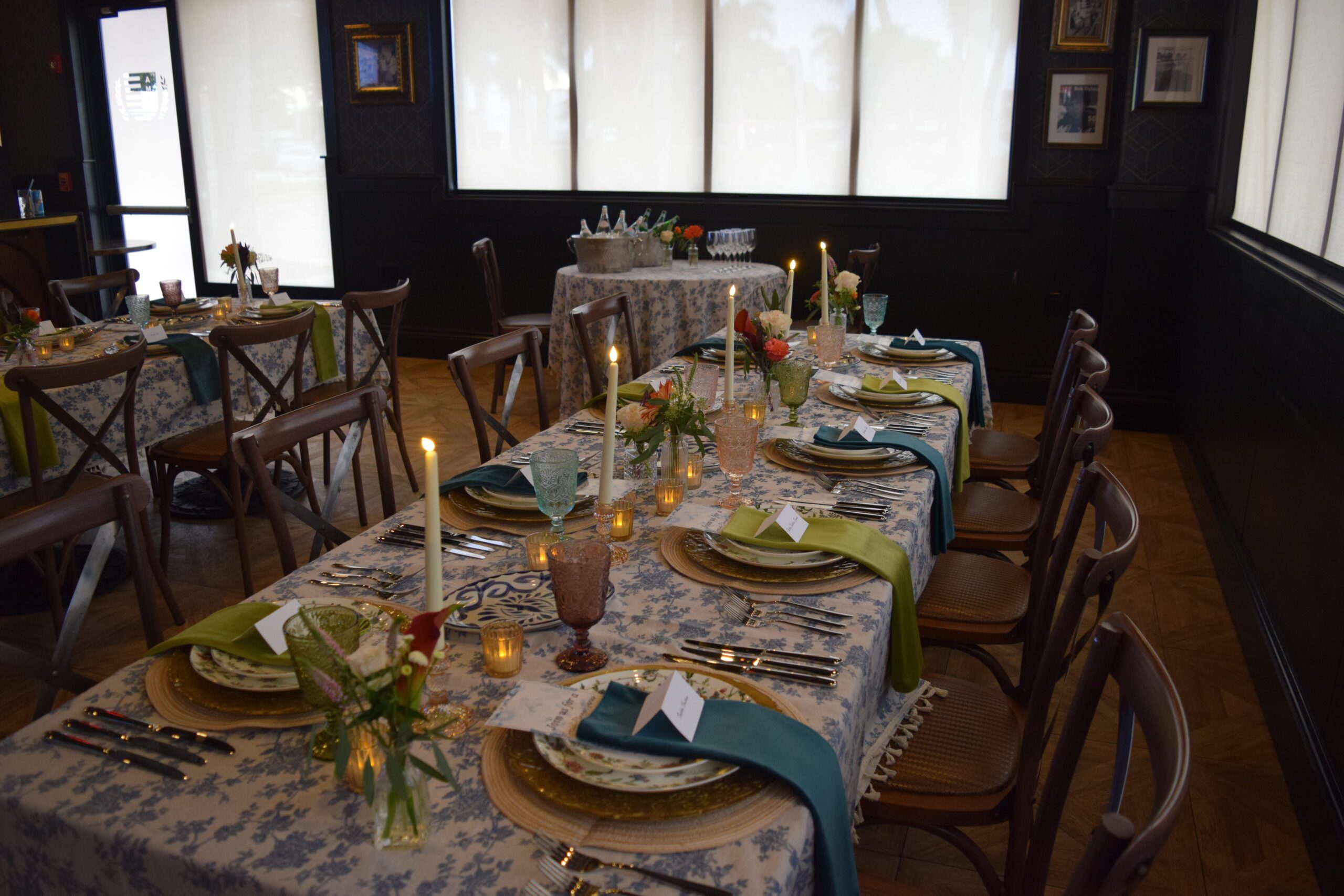 A long dining table set for a formal Bubbles & Bites meal features floral tablecloths, candles, glassware, plates, green and blue napkins, and small flower arrangements, surrounded by wooden chairs in a softly lit room.