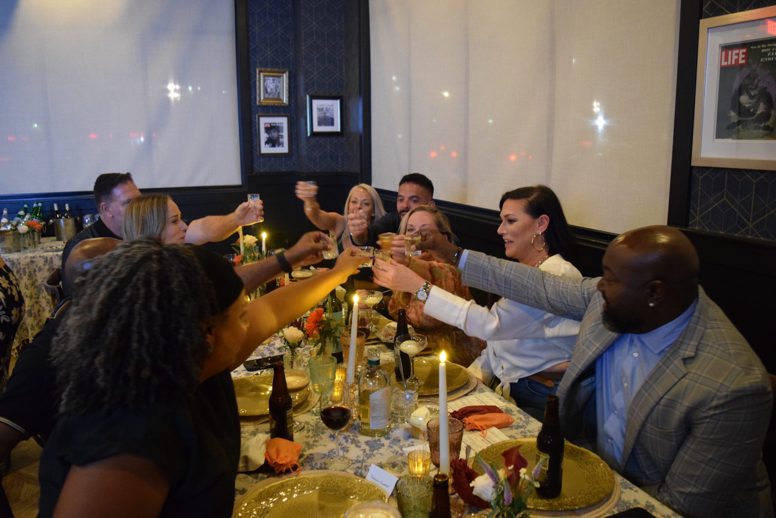 A group of people sit around a decorated dinner table, raising their glasses together in a toast. Candles, flowers, and plates set the scene as everyone smiles and celebrates, enjoying the festive Bubbles & Bites atmosphere.