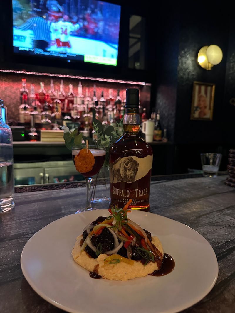 A plate of food with vegetables sits on a bar counter next to a bottle of Buffalo Trace bourbon, a cocktail from Bubbles & Bites, and a small vase with greenery. A TV shows a hockey game above a backlit bar with bottles and glasses.
