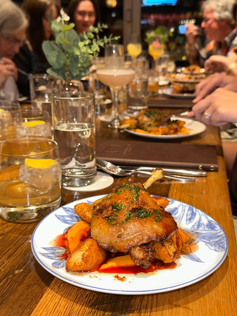 A plate with roasted chicken and potatoes sits on a wooden table surrounded by drinks and people dining together in the lively atmosphere of Bubbles & Bites restaurant.