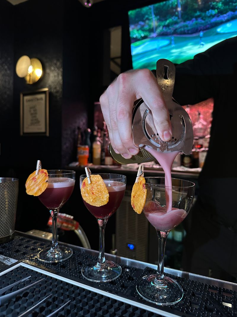 A bartender pours a pink cocktail into a coupe glass at Bubbles & Bites, with two finished cocktails beside it, each garnished with a potato chip, on the bar counter. A TV and framed picture can be seen in the background.