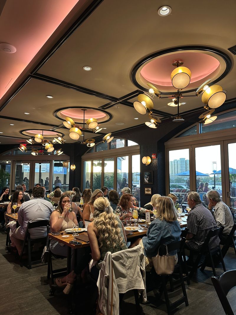 A busy Bubbles & Bites restaurant dining area filled with people eating and talking at tables under modern circular ceiling lights, with large windows revealing city buildings and an evening sky outside.