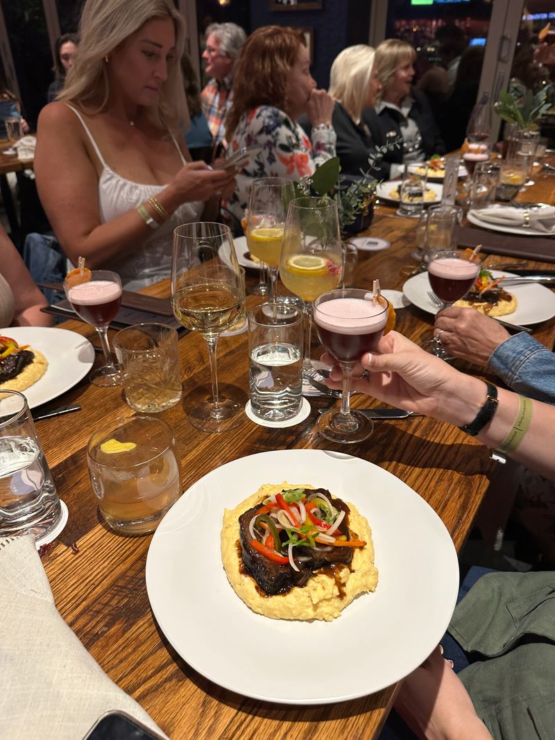 A group of people dine at Bubbles & Bites, some chatting and others on their phones. A plate of food with vegetables and sauce sits on the table, surrounded by cocktails, wine, water, and other tempting drinks.