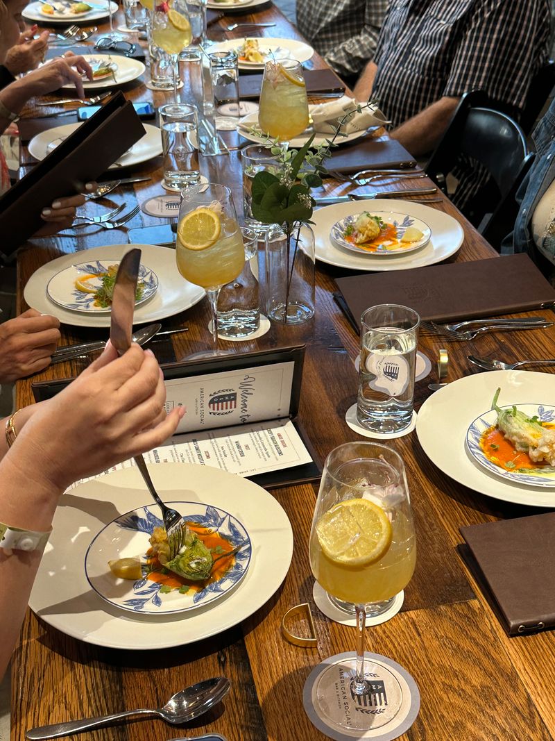A group of people sit at a wooden table, enjoying Bubbles & Bites—drinks with lemon slices and small plated appetizers. Menus, cutlery, and glasses are neatly arranged as hands reach for food in this lively, social atmosphere.