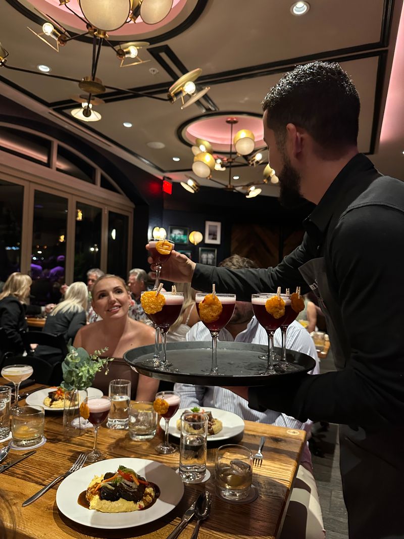 A waiter serves shrimp cocktail in martini glasses at Bubbles & Bites, a warmly lit, busy restaurant. A smiling woman in a white top looks up as he places a glass on the wooden table set with dishes and drinks.