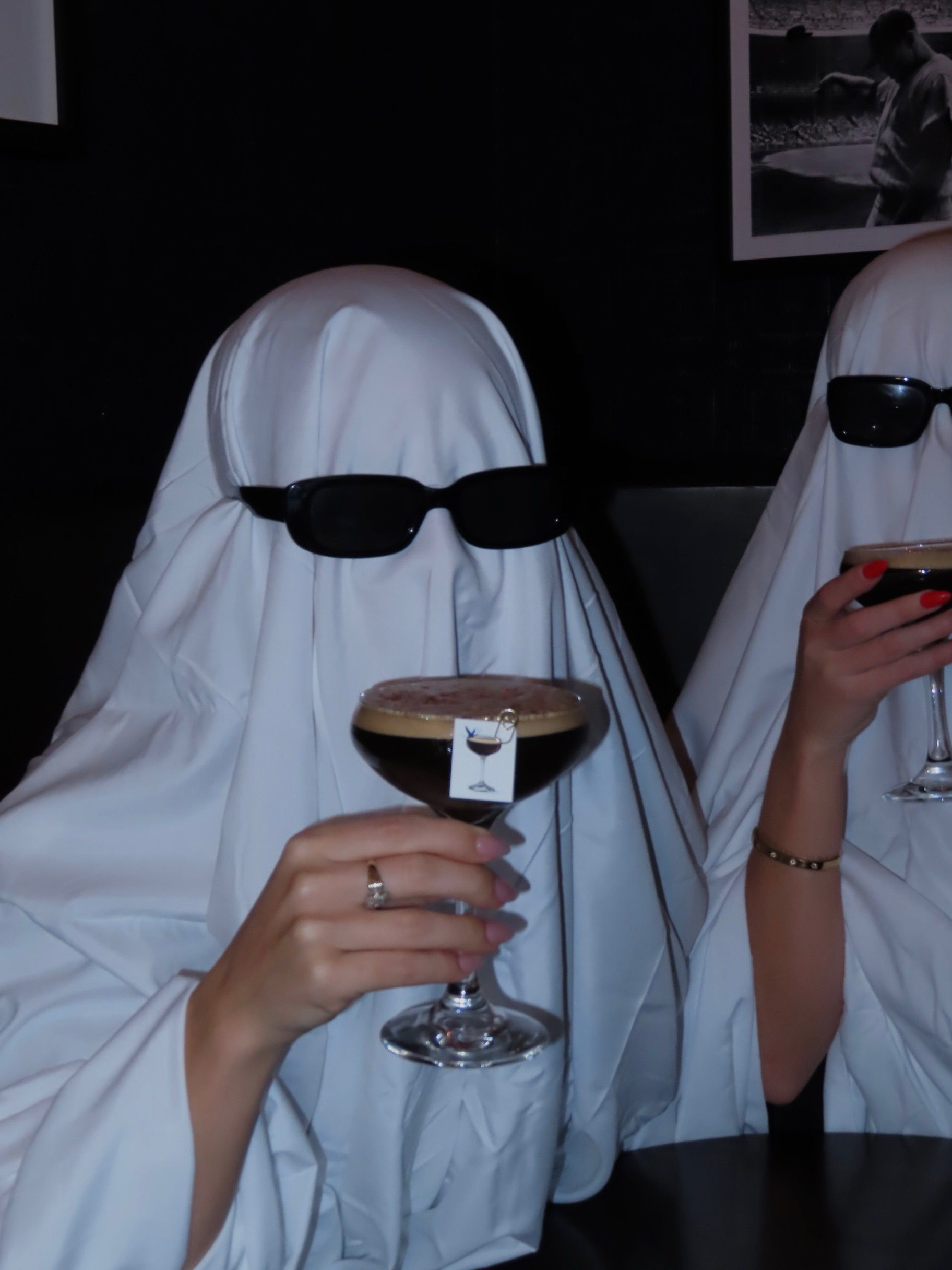Two people dressed as ghosts with white sheets and black sunglasses sit together at a dimly lit bar, each holding a cocktail glass with garnishes—capturing the playful spirit of halloween events. One person’s hand with a ring is visible.