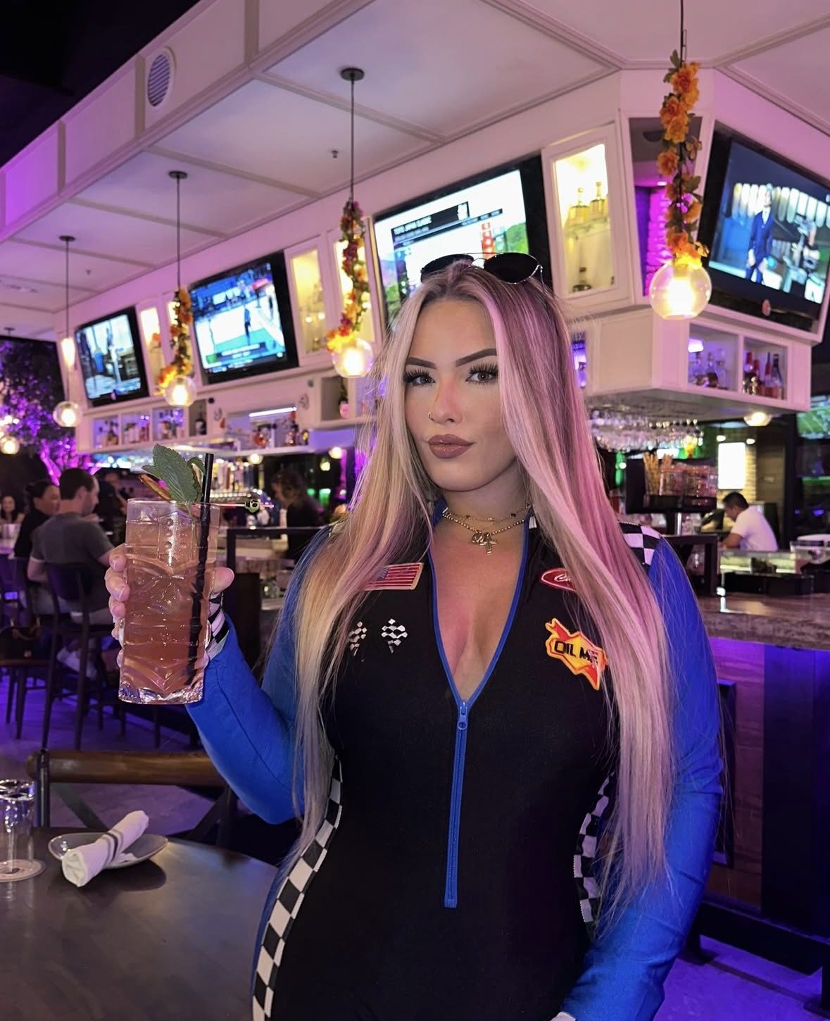 A woman with long blonde hair in a racing jumpsuit holds a cocktail with a mint garnish at a lively bar, decorated for Halloween events, with multiple TV screens and colorful lighting.