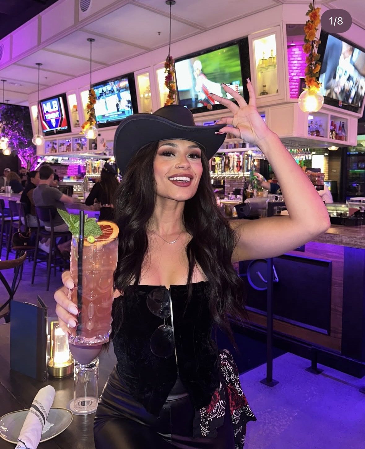 A smiling woman in a black outfit and cowboy hat holds a tall pink cocktail with garnishes at a lively bar, decorated for halloween events, with TVs and colorful lighting.