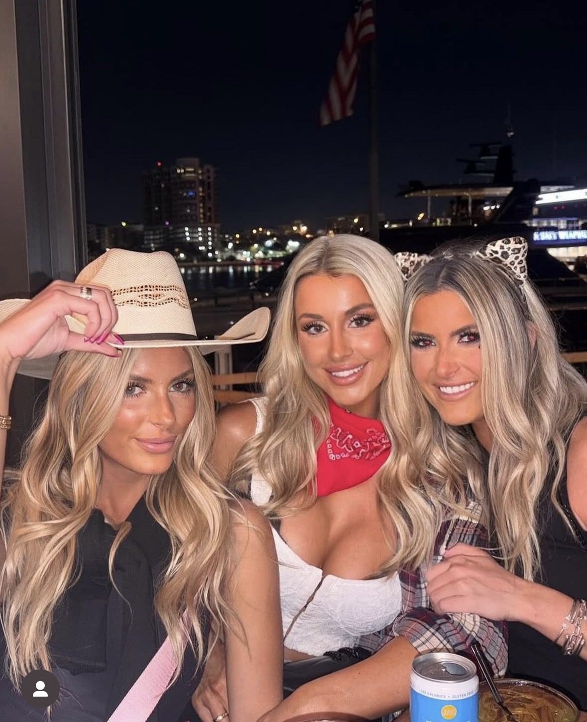 Three women with long blonde hair pose and smile at night during halloween events. One wears a cowboy hat, another a red bandana and white top, and the third has cat ears. City lights and an American flag glow in the background.