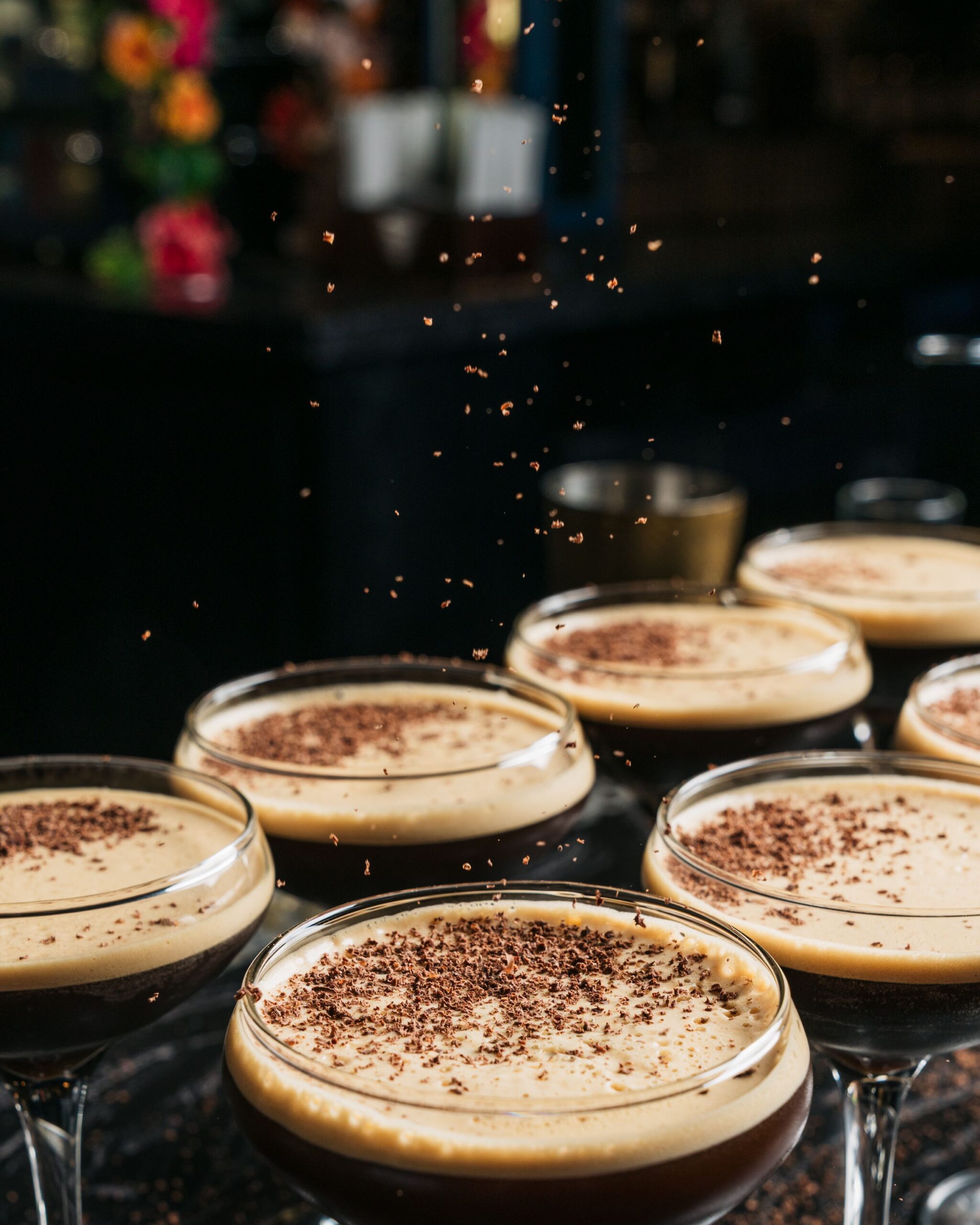Several cocktail glasses filled with frothy espresso martinis are topped with grated chocolate, as shavings fall onto the drinks. The dimly lit, slightly out-of-focus background sets a cozy mood—perfect for Wine Wednesdays indulgence.