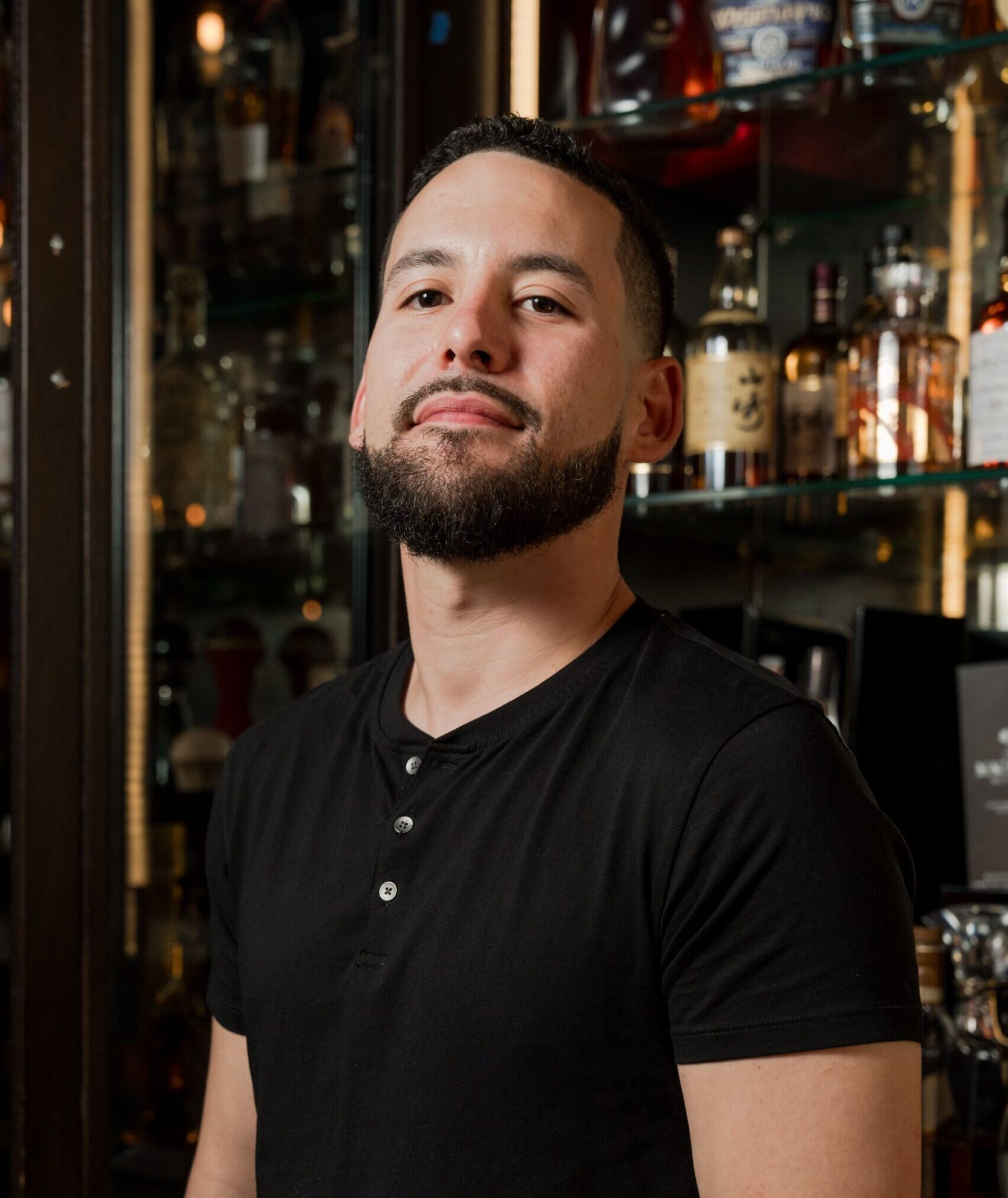 A man with a short beard and mustache, wearing a black henley shirt, stands in front of shelves filled with liquor bottles, looking confidently at the camera.